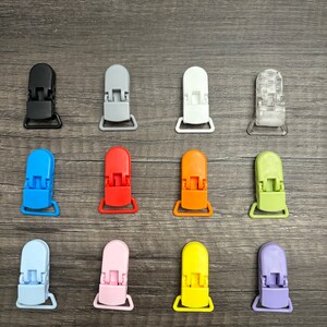 May include: Assortment of 12 plastic baby pacifier clips in various colors, including black, gray, white, blue, red, orange, green, light blue, pink, yellow, purple, and clear.
