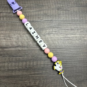 May include: A purple and yellow baby teether with a white clip and a white bead with the name "LAINEY" spelled out. The teether has a yellow, purple, and pink silicone beads and a charm of a cartoon character from Beauty and the Beast.