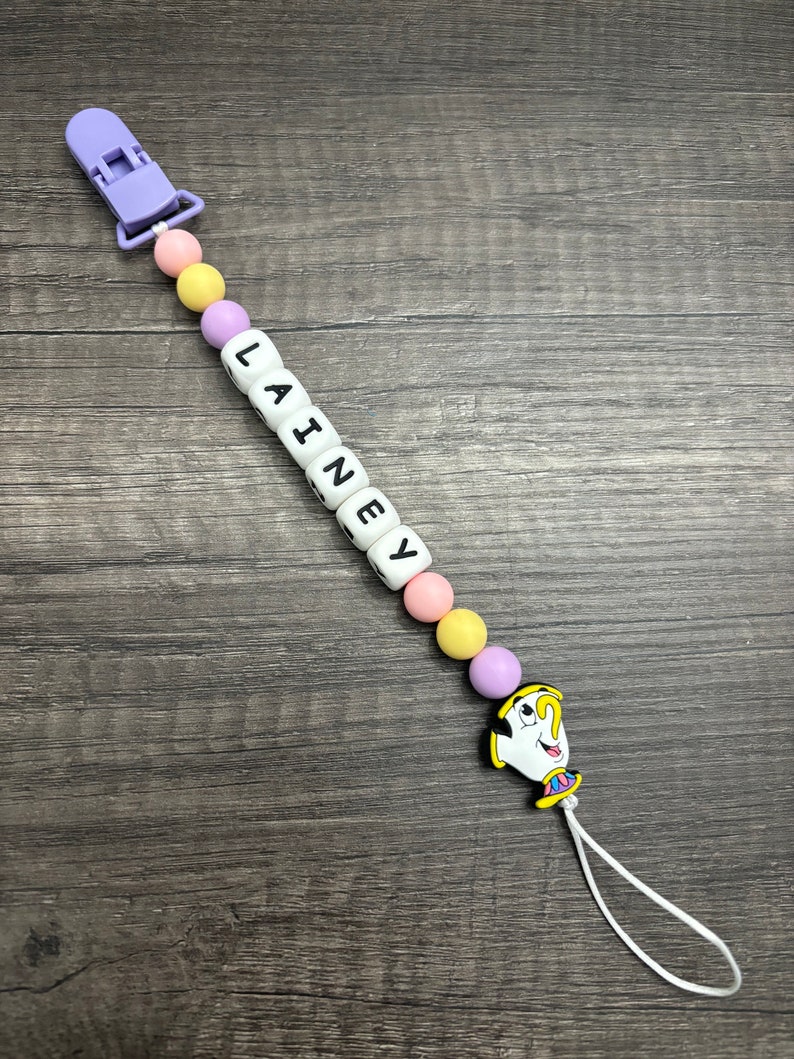 May include: A purple and yellow baby teether with a white clip and a white bead with the name "LAINEY" spelled out. The teether has a yellow, purple, and pink silicone beads and a charm of a cartoon character from Beauty and the Beast.