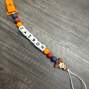 May include: A colorful silicone baby teether with the name "JAIRON" spelled out in white beads. The teether has a clip on one end and a toy character on the other end. The toy character is a cartoon cowboy with a brown hat and a yellow shirt.