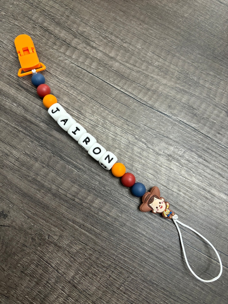 May include: A colorful silicone baby teether with the name "JAIRON" spelled out in white beads. The teether has a clip on one end and a toy character on the other end. The toy character is a cartoon cowboy with a brown hat and a yellow shirt.