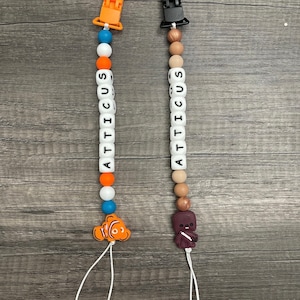 May include: Two personalized baby teethers with clips. One teether has orange, blue, and white beads with a Finding Nemo character. The other teether has brown, white, and beige beads with a Star Wars character.