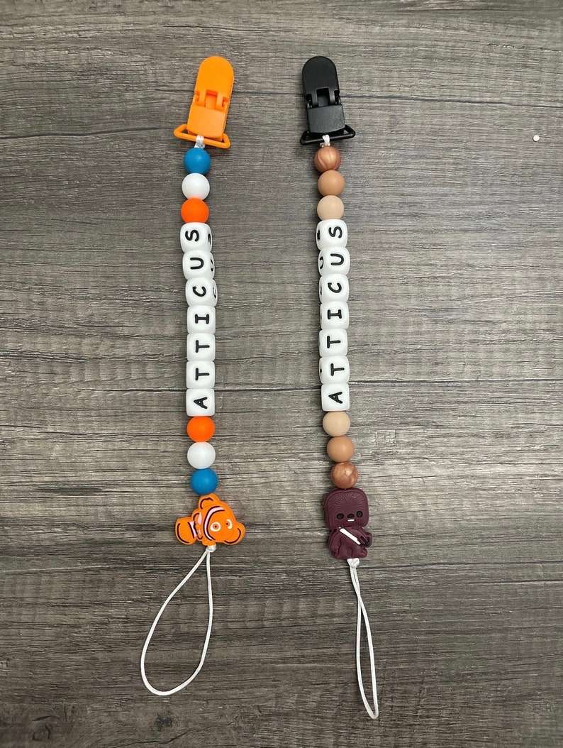 May include: Two personalized baby teethers with clips. One teether has orange, blue, and white beads with a Finding Nemo character. The other teether has brown, white, and beige beads with a Star Wars character.