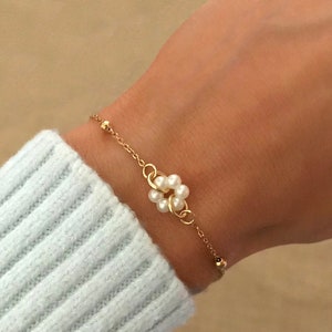 Könnte beinhalten: Ein goldenes Kettenarmband mit einem Perlenblumen-Anhänger. Das Armband befindet sich am Handgelenk einer Person.