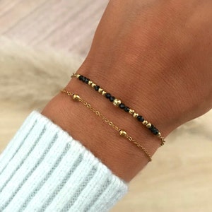 Peut inclure: Deux bracelets dorés sont présentés sur un poignet. L'un comporte de petites perles noires et dorées, tandis que l'autre est une chaîne délicate avec de petites perles dorées. Les bracelets sont superposés.