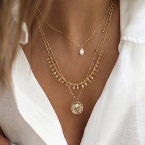 Peut inclure: Un collier en or à plusieurs rangs avec un pendentif en perle délicate et un pendentif en disque texturé plus grand. Le collier est porté par une personne avec une chemise blanche.