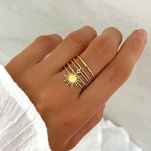 May include: Gold ring set featuring a sun charm. The rings are stacked on a finger and are made of thin, delicate metal.
