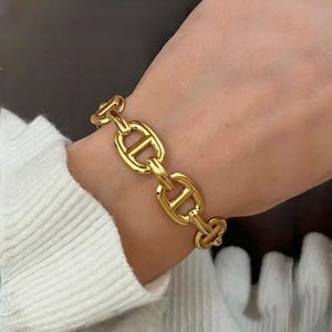 Peut inclure: Un bracelet chaîne doré avec une série de maillons ovales entrelacés. Le bracelet est porté au poignet et les maillons ont un design distinctif. Le bracelet est de couleur or brillant et semble être en métal.