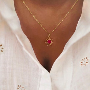 Może przedstawiać: Złoty łańcuszek z czerwonym słonecznym wisiorem. Słońce ma złotą obwódkę i czerwony środek.