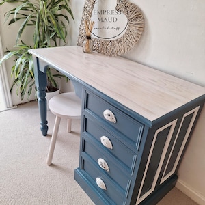 May include: A desk with a light wood-toned top and a dark blue base featuring four drawers with white shell-shaped pulls. The side of the desk has white rectangular trim detailing. A small, light-coloured wooden stool is tucked underneath the desk.