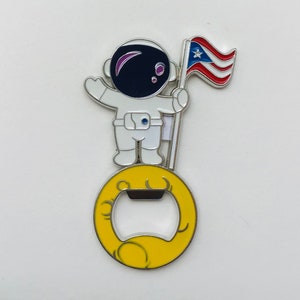 May include: A bottle opener featuring a white astronaut figure holding a Puerto Rican flag. The astronaut stands on a yellow moon-shaped base with a silver bottle opener. The design has a colorful enamel finish.