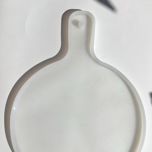 May include: A white, round silicone mold with a handle. The mold has a circular base with a raised edge and a handle with a small hole at the top. The object is designed for crafting or food preparation.