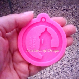 May include: Pink silicone mold for resin casting, featuring a design of a building with the text "ArtelsieCraftDesign" and "Angel Wings" on the mold. The mold is 2.5 inches in diameter.