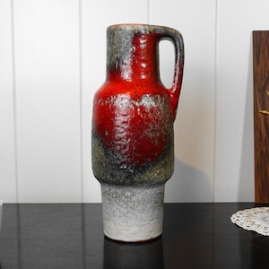 Può includere: Un vaso in ceramica rosso e nero con una superficie testurizzata e un manico. Il vaso ha una base bianca ed è alto circa 25 cm.