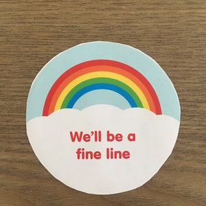 May include: A round sticker featuring a rainbow and clouds. The text "We'll be a fine line" is written in red.