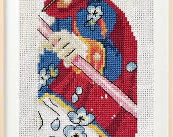 May include: Cross-stitch embroidery of a person wearing a red and blue kimono with white flowers. The person is holding a pink stick. The embroidery is framed in a light wood frame.