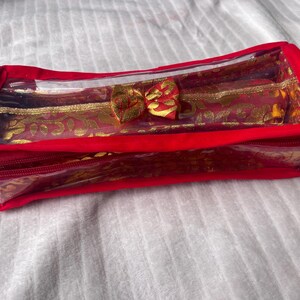 May include: A rectangular, red cosmetic bag with a clear plastic top and a red zip. The bag features a gold and red patterned interior and a decorative bow. The bag is designed for storing makeup and beauty products.