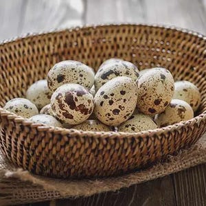 May include: A woven basket filled with speckled quail eggs. The eggs are a creamy white color with brown spots and are nestled on a burlap cloth. The basket is made of a light brown woven material and sits on a wooden surface.