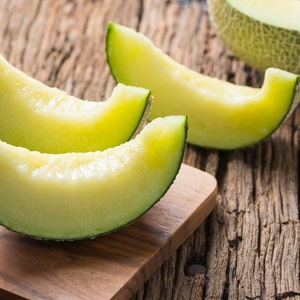 May include: Three slices of green melon on a wooden cutting board. The melon is light green and has a white flesh. The cutting board is brown and has a rough texture.