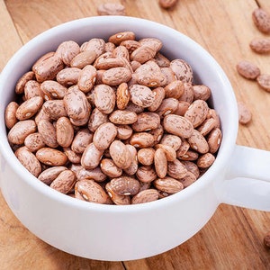 May include: A white ceramic bowl filled with pinto beans. The beans are light brown with darker brown speckles. The bowl has a handle on each side. The bowl is sitting on a wooden surface.