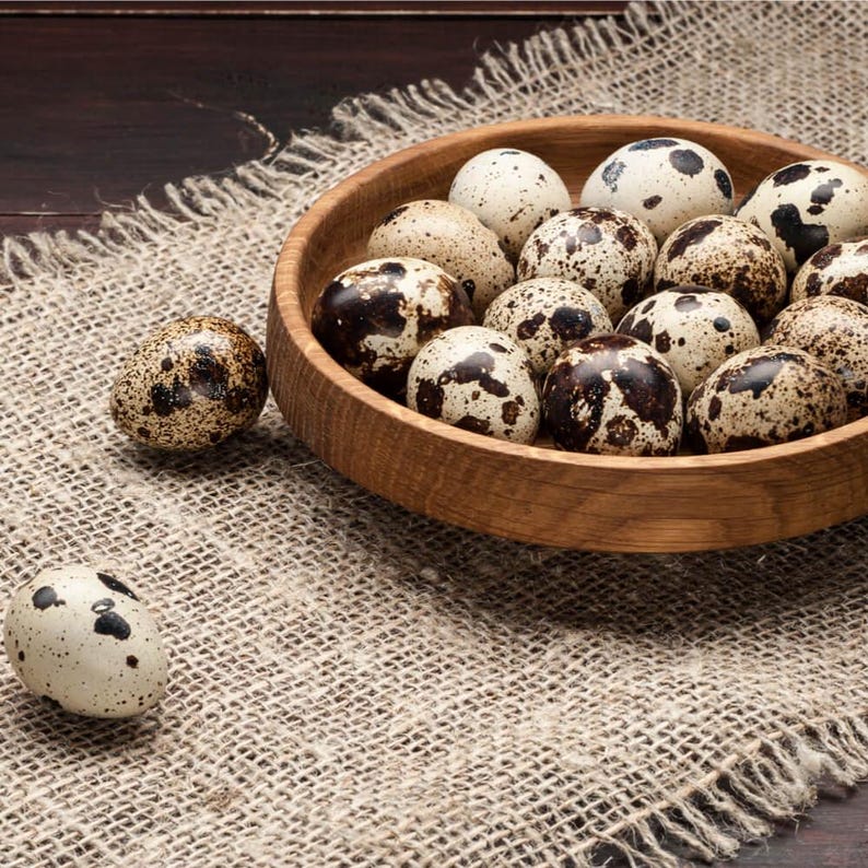 May include: A wooden bowl filled with speckled quail eggs, resting on a burlap cloth. The eggs have a creamy white base with brown spots. The bowl is made of light brown wood. One egg is isolated on the cloth.