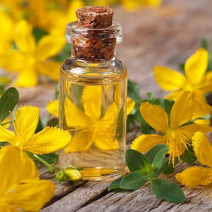 May include: A small glass bottle with a cork stopper filled with a clear yellow liquid. The bottle is surrounded by yellow flowers with green leaves on a wooden surface.