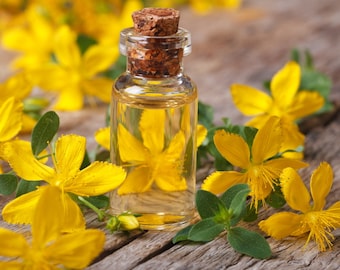 May include: A small glass bottle with a cork stopper filled with a clear yellow liquid. The bottle is surrounded by yellow flowers with green leaves on a wooden surface.