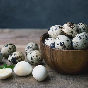 May include: A wooden bowl filled with speckled quail eggs, alongside several whole and halved eggs on a wooden surface. The eggs have a creamy white base with dark brown spots. A sprig of green mint is also present.