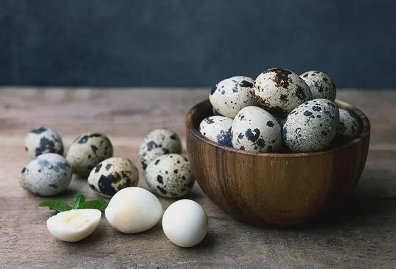 May include: A wooden bowl filled with speckled quail eggs, alongside several whole and halved eggs on a wooden surface. The eggs have a creamy white base with dark brown spots. A sprig of green mint is also present.