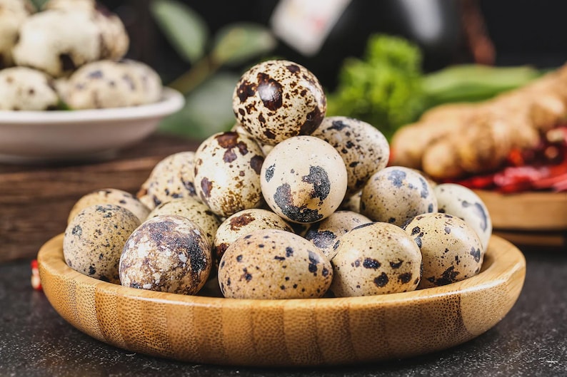 May include: A wooden bowl filled with speckled quail eggs. The eggs are a creamy beige color with brown and black spots. Other eggs are in a white bowl in the background. Fresh ingredients are in the background.