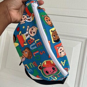 May include: A blue fanny pack with a white zipper and black strap. The pack is covered in a colorful pattern featuring cartoon characters and the text "LET'S BUILD". The bag is held by a hand.