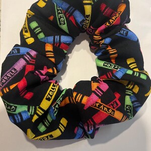 May include: A black scrunchie with a colorful crayon pattern. The fabric features various crayon colors, including yellow, green, orange, purple, and blue, with the crayon names printed on them. The scrunchie is designed for hair styling.