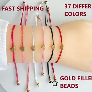 May include: Seven colorful string bracelets with gold heart charms and gold beads. The bracelets are tied around a wrist. The text "FAST SHIPPING" and "37 DIFFERENT COLORS" is at the top of the image. The text "GOLD FILLED BEADS" is at the bottom of the image.
