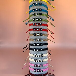 May include: A display of personalized macrame bracelets in various colors, each featuring letter beads and a heart bead. The bracelets are arranged on a gray cylindrical stand against a peach background. The bracelets are in shades of teal, yellow, blue, green, red, and black.