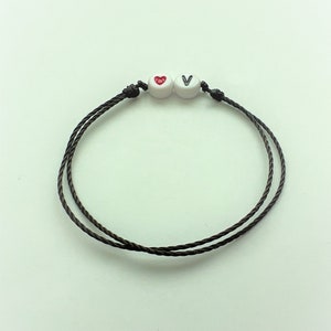 Könnte beinhalten: Ein braunes Schnurarmband mit einer weißen Perle mit einem roten Herzen und einer weißen Perle mit dem Buchstaben V.