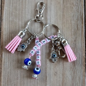 Könnte beinhalten: Rosa-blauer Schlüsselanhänger mit dem Namen "SERAH", der mit Perlen ausgeschrieben ist, zwei blauen bösen Augen und zwei silbernen Hamsa-Anhängern mit blauen bösen Augen. Der Schlüsselanhänger hat einen silbernen Karabinerhaken und zwei silberne Schlüsselringe.