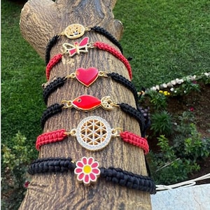 Pode incluir: Um conjunto de seis pulseiras de corda vermelha e preta com diferentes pingentes dourados. Os pingentes incluem um sol, uma borboleta, um coração, um peixe, uma flor da vida e uma flor.