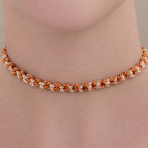 Może przedstawiać: Naszyjnik typu choker wykonany z plecionego sznurka w naprzemiennych odcieniach spalonej pomarańczy i kremu. Tkany wzór charakteryzuje się powtarzalnym splotem węzłowym, który nadaje mu teksturowany, cylindryczny wygląd wokół szyi.