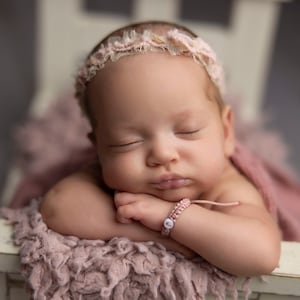 May include: A sleeping baby girl wearing a white lace headband and a pink bracelet with a white letter charm. The baby is lying on a pink fuzzy blanket.