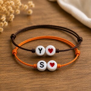 Pode incluir: Duas pulseiras de cordão ajustável, uma castanho-escura e uma laranja, cada uma com duas contas redondas brancas. A pulseira castanha apresenta a letra Y e um coração vermelho, enquanto a pulseira laranja apresenta a letra S e um coração vermelho.