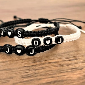 Puede incluir: Dos pulseras de amistad con hilo negro y blanco, con las letras J, S, D y un dije de corazón.
