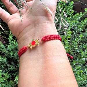 Puede incluir: Pulsera de cuerda roja con un dije de tortuga dorada. La tortuga tiene un centro rojo y está unida a la pulsera con un anillo dorado.