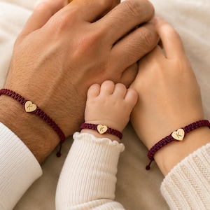 May include: Three matching adjustable bracelets featuring a deep burgundy woven cord and a small gold-toned heart-shaped charm. Each charm is engraved with the letter K in a simple serif font. The bracelets are designed for different wrist sizes.
