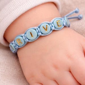 Peut inclure: Un bracelet bleu clair avec les lettres "RIVE" en or. Le bracelet est composé de perles rondes bleu clair avec des lettres dorées, enfilées sur un cordon bleu clair. Le bracelet est porté au poignet d'un bébé.