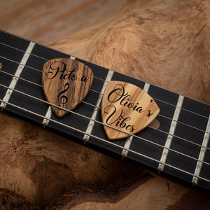 Puede incluir: Dos púas de guitarra de madera con texto grabado personalizado. La primera púa dice "Pick a" con un símbolo de clave de sol. La segunda púa dice "Olivia's Vibes".