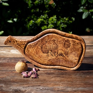 Può includere: Un tagliere in legno con la scritta "Sunday Roast". Il tagliere è in legno d'ulivo e ha un manico. Un bulbo d'aglio e diversi spicchi sono accanto al tagliere. Il tagliere è su una superficie di legno.