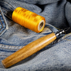 Peut inclure: Une bobine de fil jaune et une aiguille en bois avec une pointe métallique, posées sur un tissu en jean bleu.