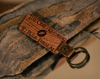 May include: Wooden keychain with the text "THE ONE (KEY) RING TO RULE THEM ALL" and a ring graphic. The keychain has a leather strap and metal rings. The wood has a natural grain.