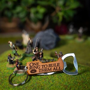 Può includere: Un apribottiglie in legno con un anello di metallo e un cavatappi. L'apribottiglie ha l'iscrizione "One Ring to Rule Them All" con un piccolo simbolo ad anello. L'apribottiglie è su una superficie verde con figure in miniatura di persone.