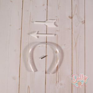 May include: White plastic arrow and arrow-shaped decorations, along with a clear plastic headband and a small black object. The items are arranged on a light wood-grain surface. A logo with the text "Dolly Pop" is in the bottom right corner.